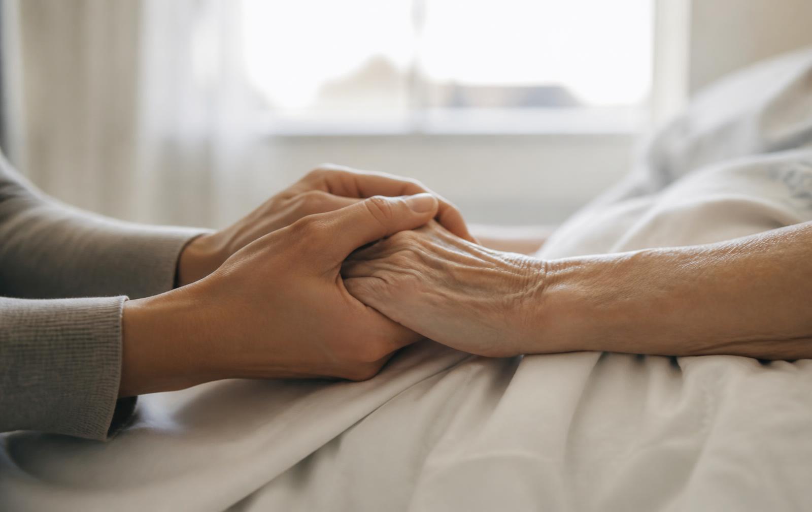 A caregiver holding the hand of an elderly patient in a hospital bed